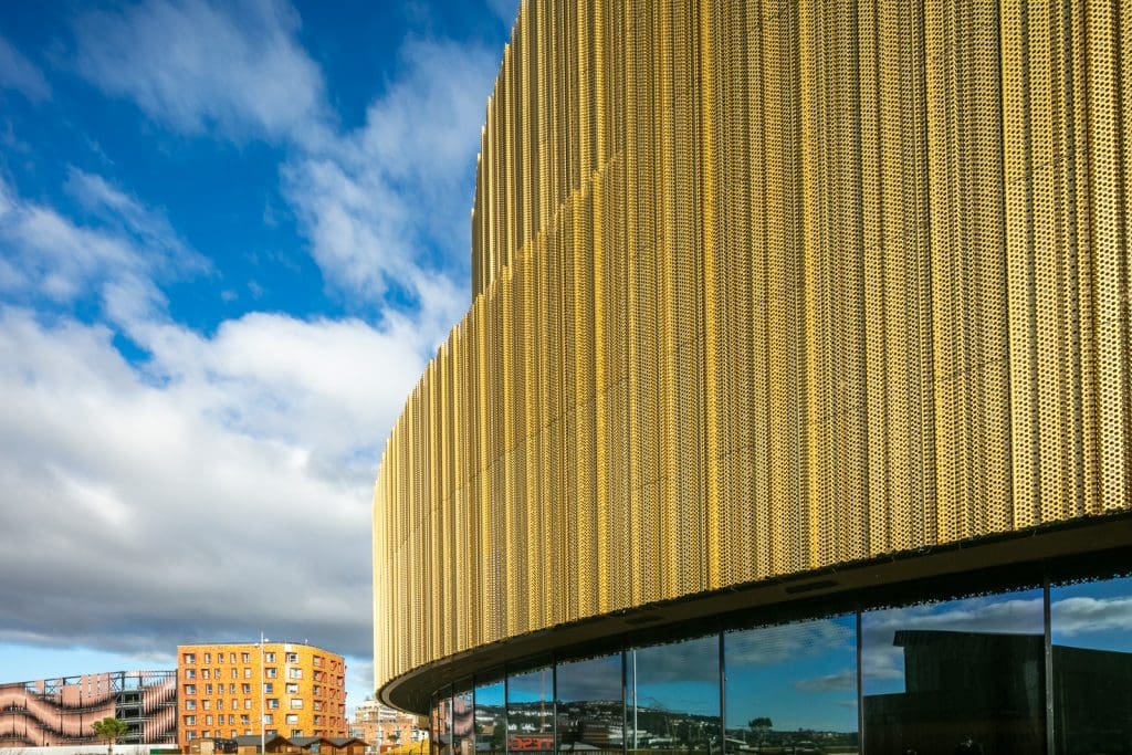 Modern building with a curved facade covered in gold perforated panels, reflecting the sky and surrounding structures in its glass windows. Blue sky with scattered clouds and a red brick building are visible in the background.