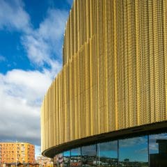 Modern building with a curved facade covered in gold perforated panels, reflecting the sky and surrounding structures in its glass windows. Blue sky with scattered clouds and a red brick building are visible in the background.