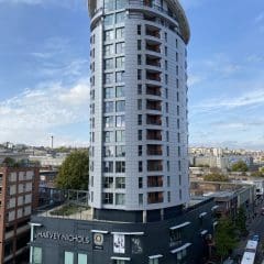 A tall, modern cylindrical building with many windows rises above a Harvey Nichols store. The sky is blue with scattered clouds, and people and vehicles move along the surrounding streets.