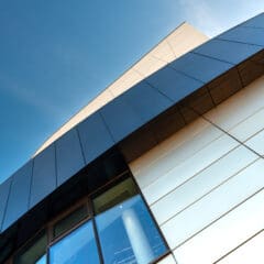 Close-up view of a modern building with reflective glass and metal panels, captured from a low angle against a clear blue sky. Geometric lines and sharp angles dominate the architecture.