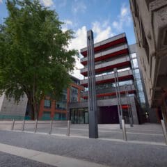 A modern building with red and black accents stands behind a tree, metal poles, and a gravel courtyard, with another concrete building on the right and a partly cloudy sky above.