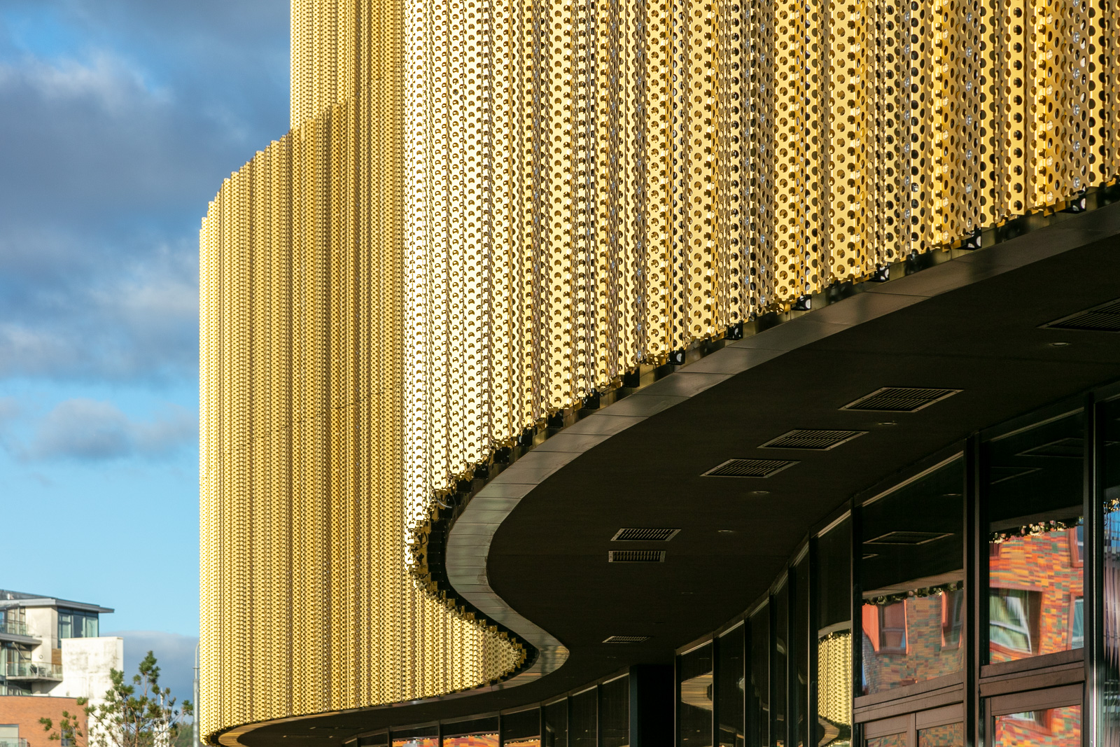 Close-up of a modern building with a curved facade covered in gold-colored, perforated metal panels, reflecting sunlight. The lower part features dark glass windows and overhangs, with part of the sky and nearby buildings visible.