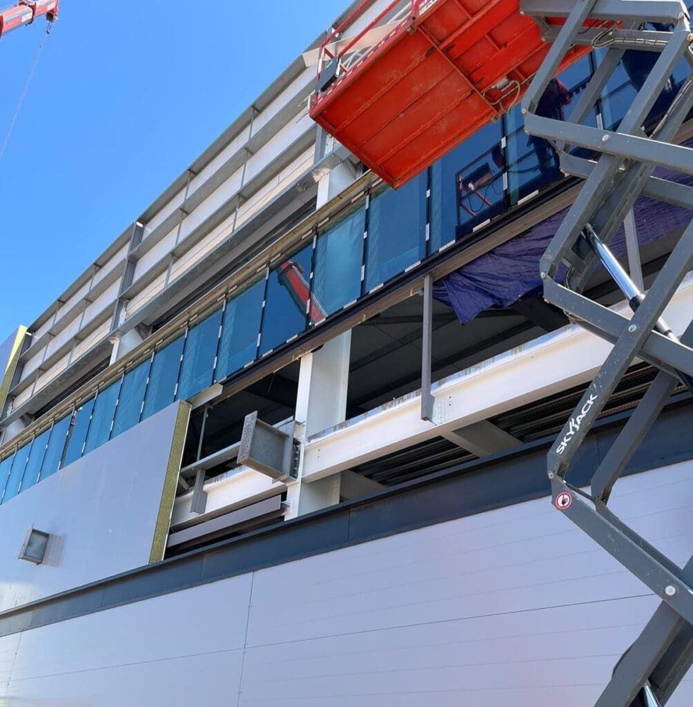 Construction site showing the exterior of a multi-story building with glass panels being installed. Two red and black scissor lifts are visible, and part of the building framework is exposed under a clear blue sky.