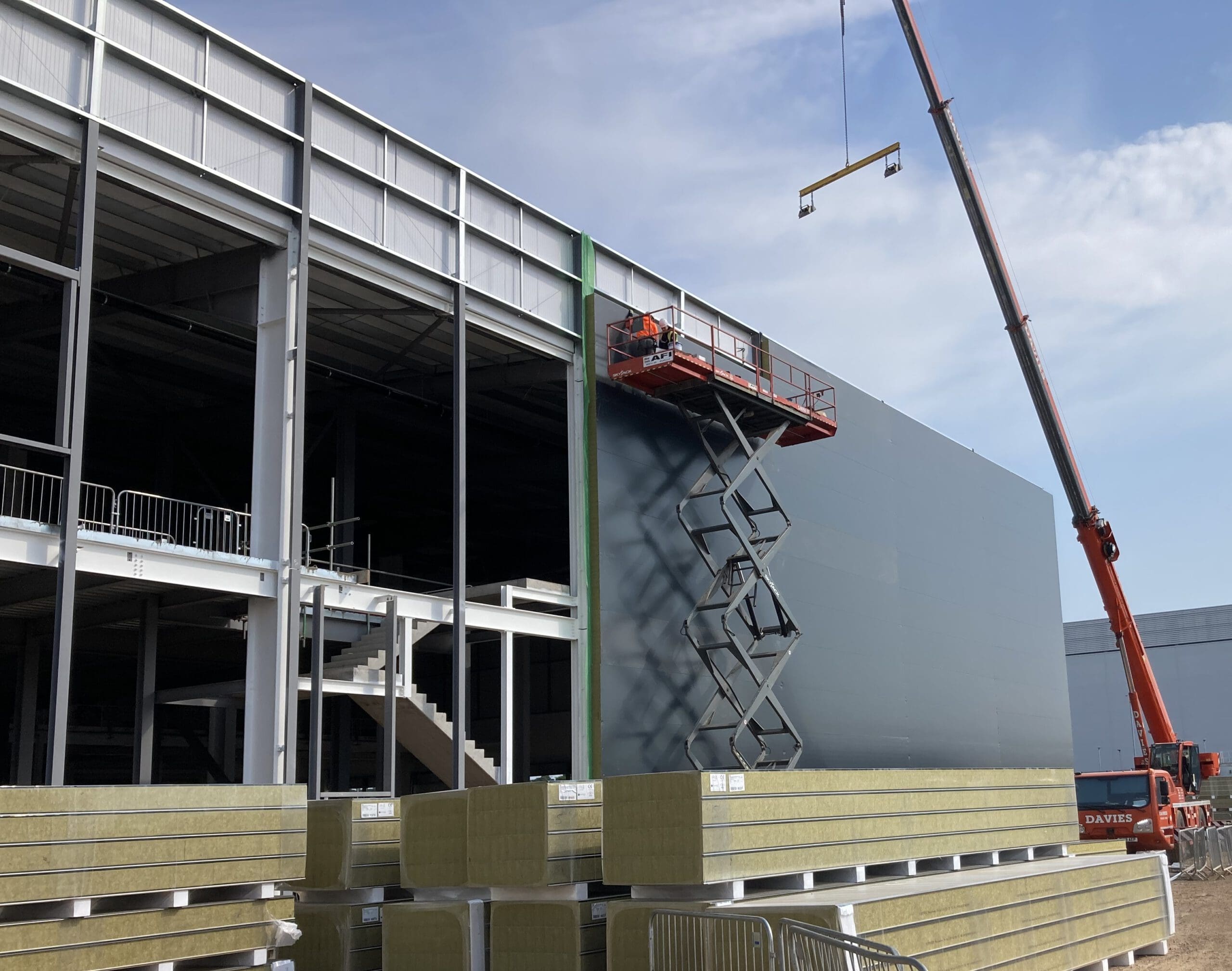 Construction site with a large crane and a worker on a scissor lift installing dark wall panels on a building exterior. Stacks of insulation materials are in the foreground under a partly cloudy sky.