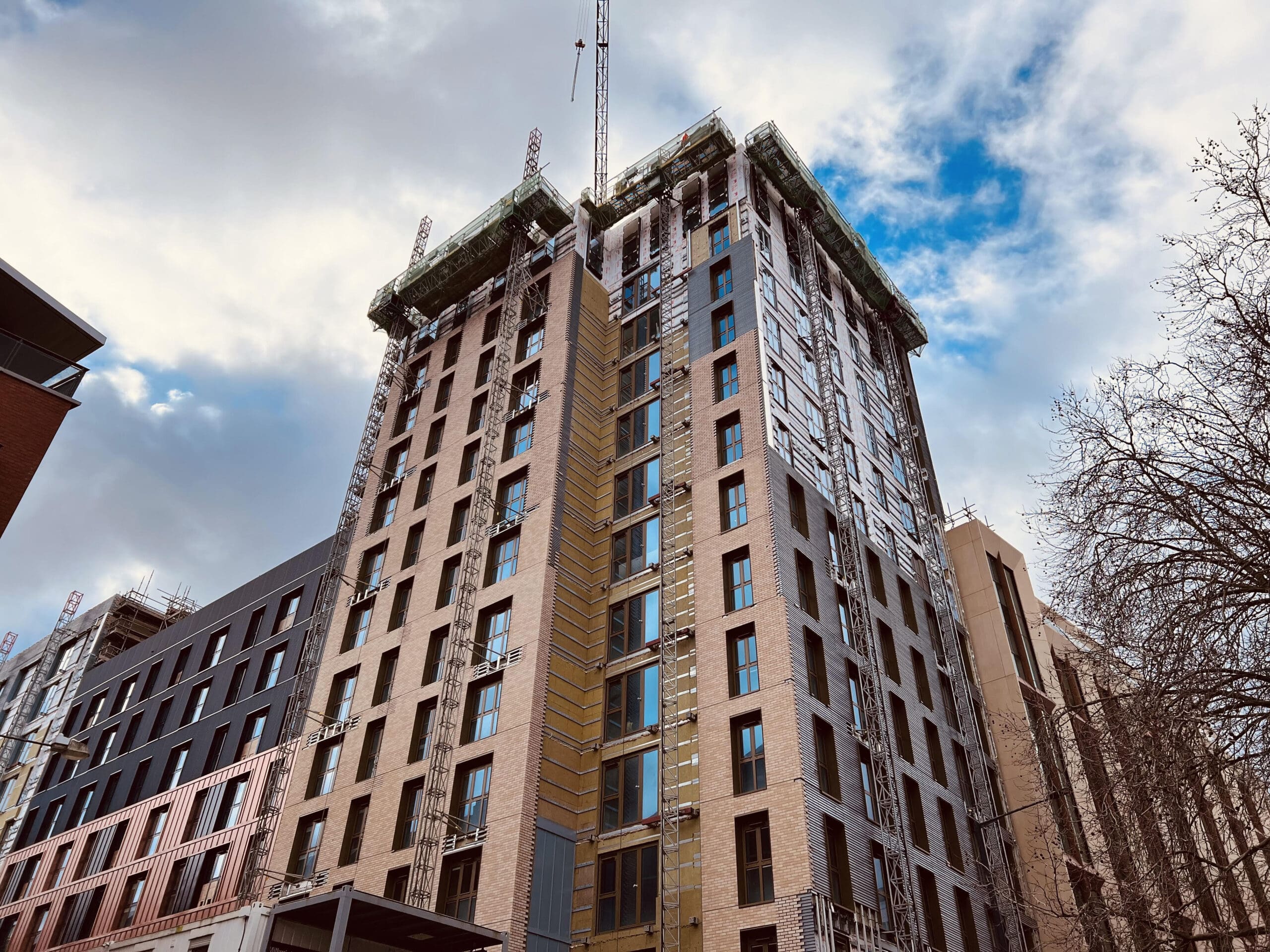 A tall, modern apartment building under construction with scaffolding and cranes, set against a partly cloudy sky. Some windows are installed, and parts of the facade are unfinished. Leafless trees are visible to the right.
