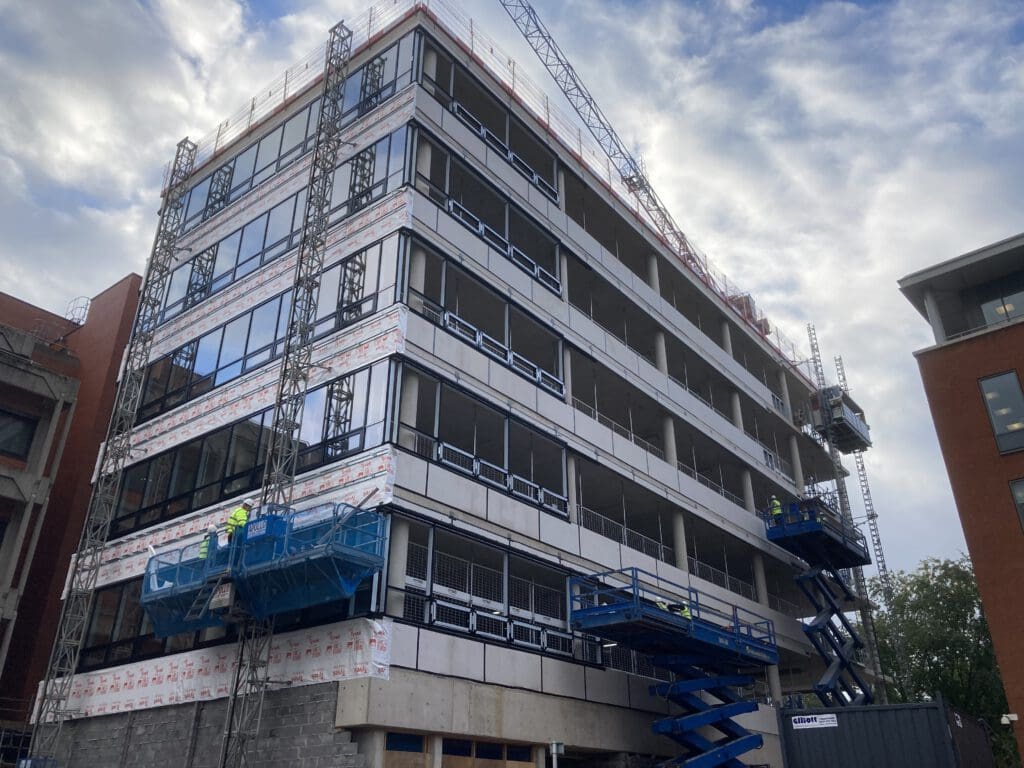 A multi-story building under construction, with workers on blue scissor lifts installing panels on the facade. The sky is cloudy, and construction cranes and scaffolding are visible around the building.