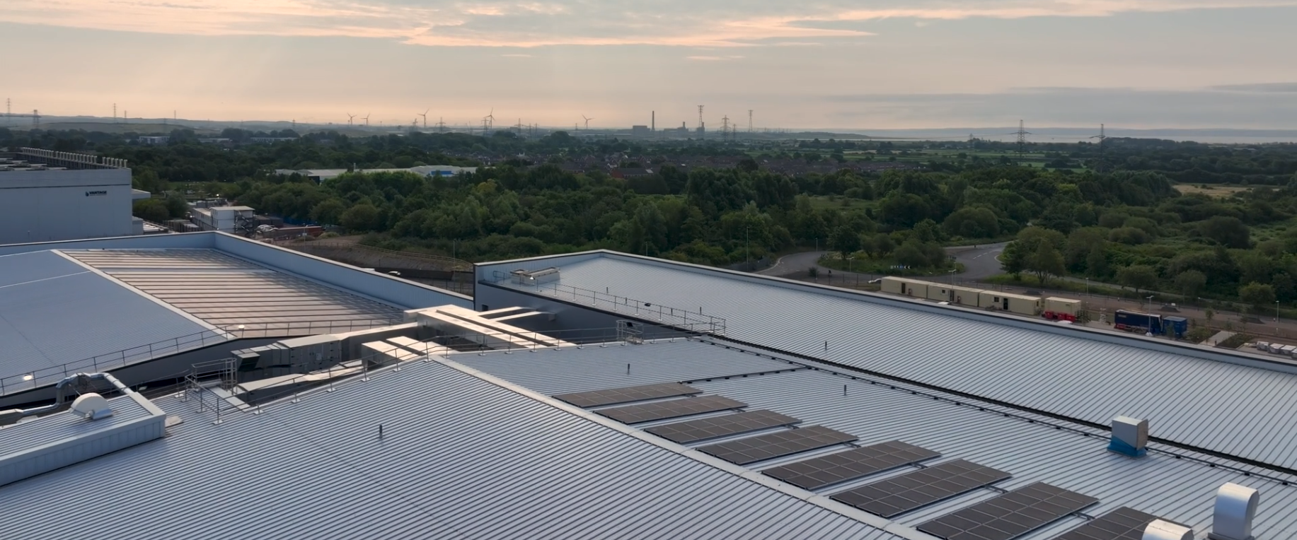 Aerial view of a large industrial building with rooftop solar panels, surrounded by green trees and distant fields under a partly cloudy sky at sunset.
