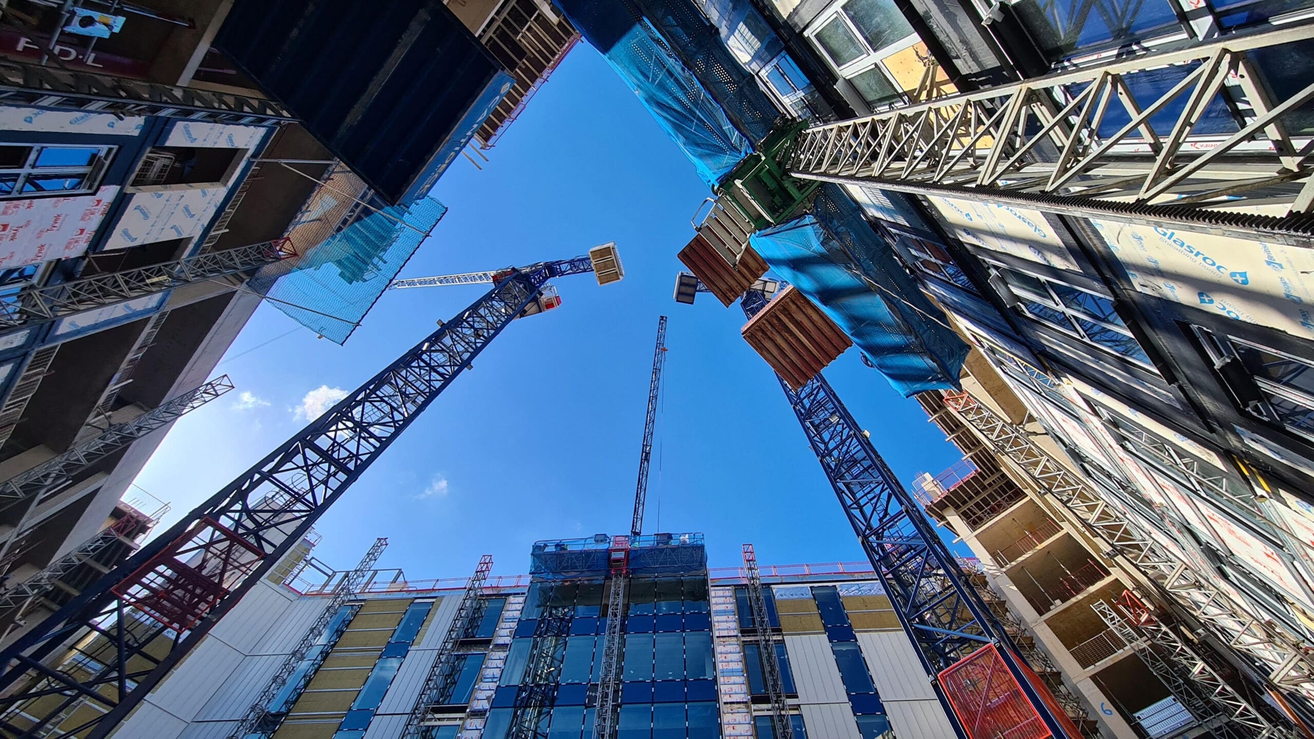 View looking up at multiple construction cranes and partially built modern buildings under a bright blue sky, with scaffolding and construction materials visible on the structures.