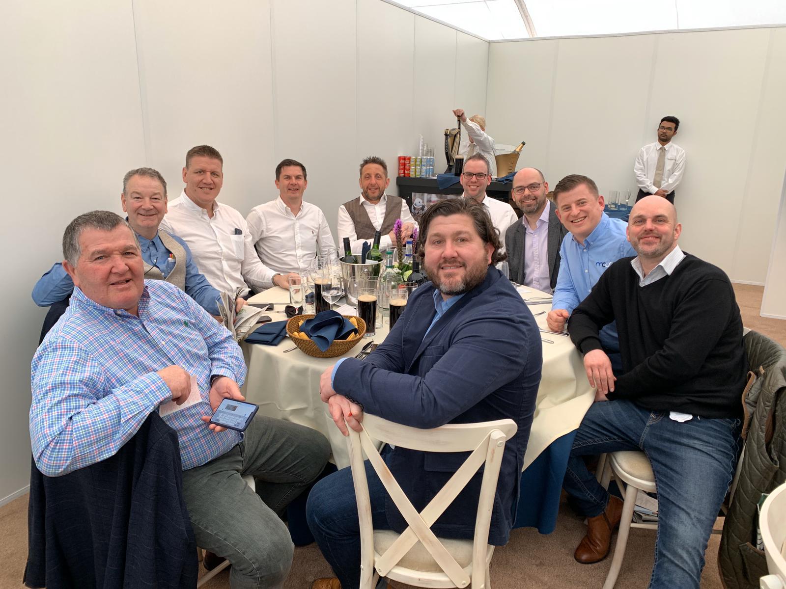 A group of eleven men sit and stand around a round table with drinks and food in a bright, white room. Most are smiling at the camera, dressed in smart-casual clothing. Two servers stand in the background.