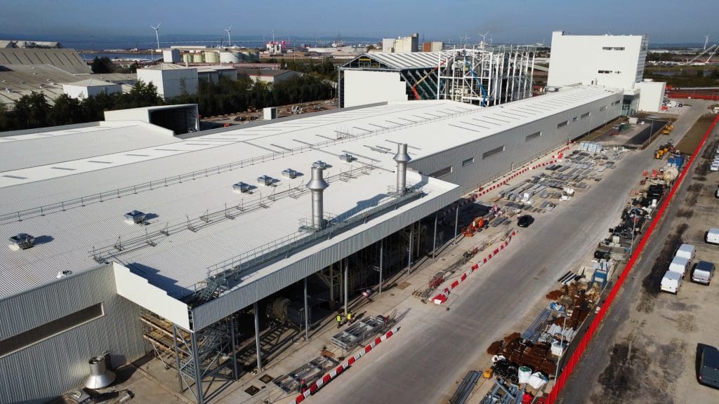 Aerial view of an industrial facility under construction, with white buildings, construction materials, vehicles, and workers along a long paved road; wind turbines and industrial structures are visible in the background.