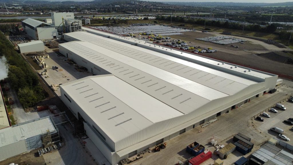 Aerial view of a large industrial warehouse surrounded by parked cars, construction materials, and green areas, with a parking lot and roads visible in the background.