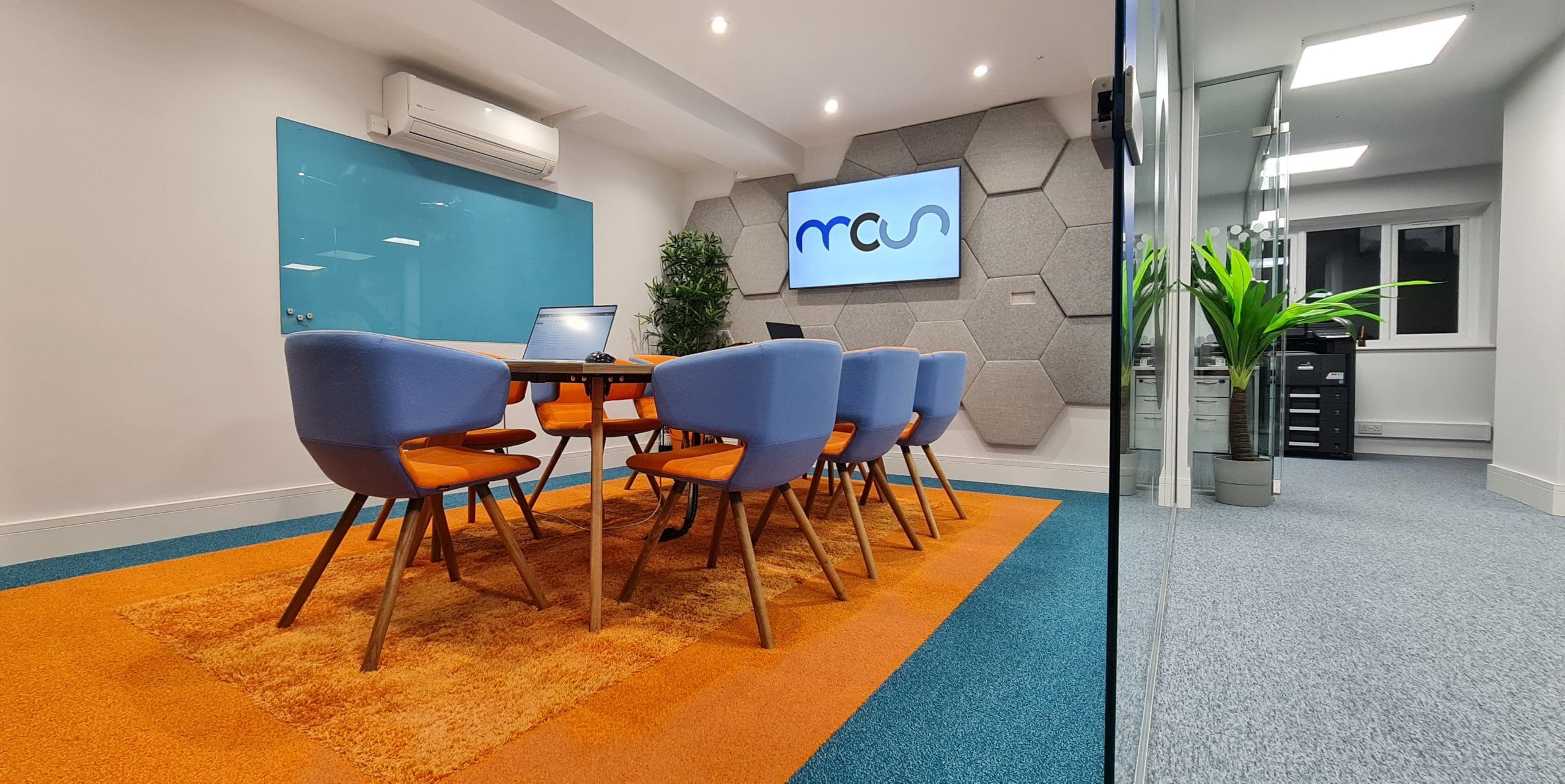 A modern conference room with a long table, eight blue and orange chairs, a wall-mounted screen, hexagonal wall panels, a blue glass board, and a bright orange rug on the floor. A potted plant is visible near the entrance.