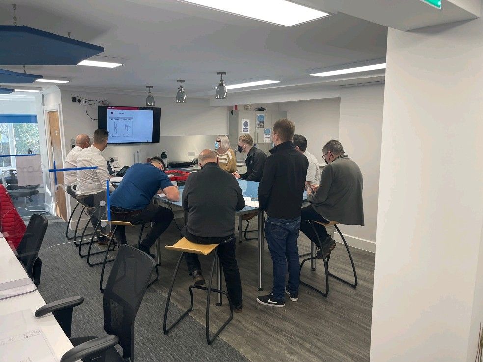 A group of people sit around a large table in a modern office, facing a screen displaying a presentation. Some are taking notes, and others are listening attentively. The room is bright with natural and artificial light.